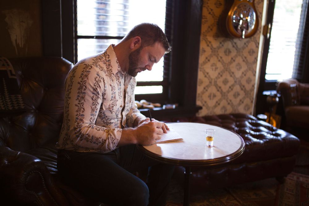 A person sits at a rustic wooden table writing or drawing in a dimly lit room with vintage decor.