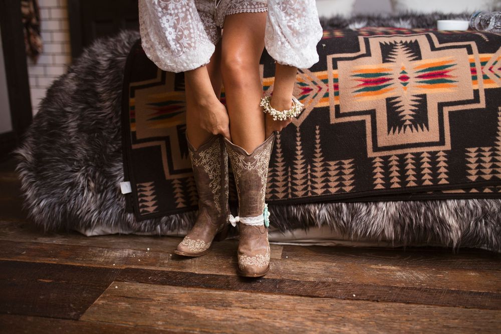 A close-up view of brown leather cowboy boots with white lace trim worn with a white dress on a wooden floor.