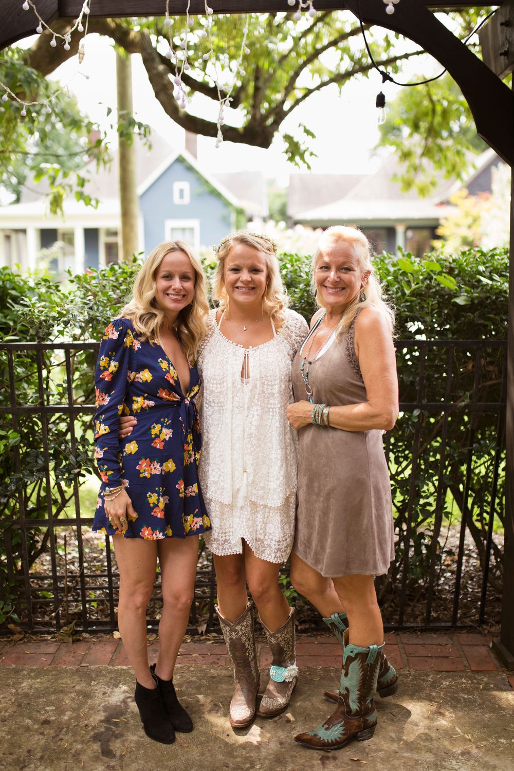 Three friends in cowboy boots and casual dresses pose together against a garden fence on a sunny day.