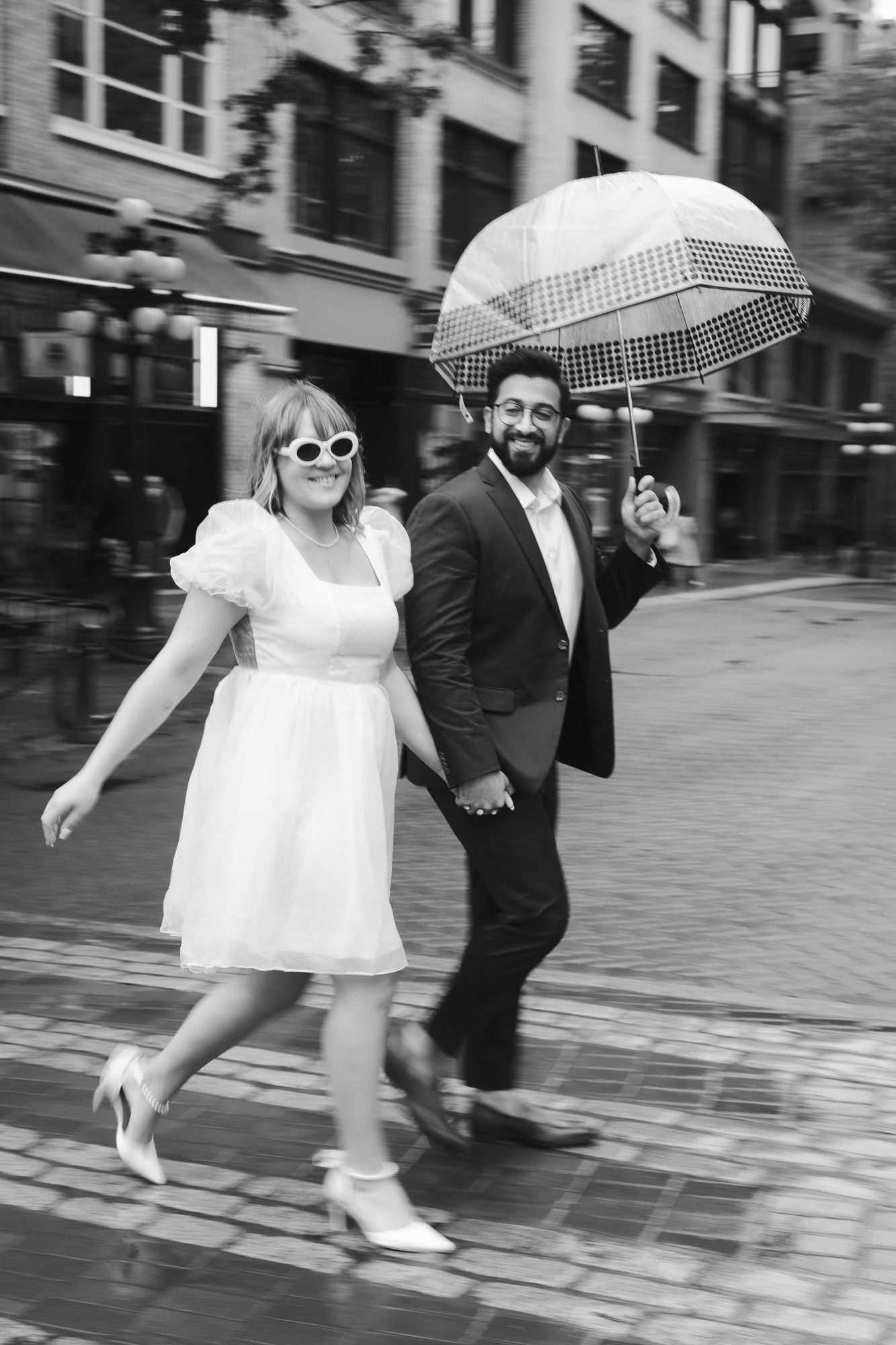 An engaged couple walks across the street in Gastown, Vancouver. She wears a white dress, he wears a black suit.