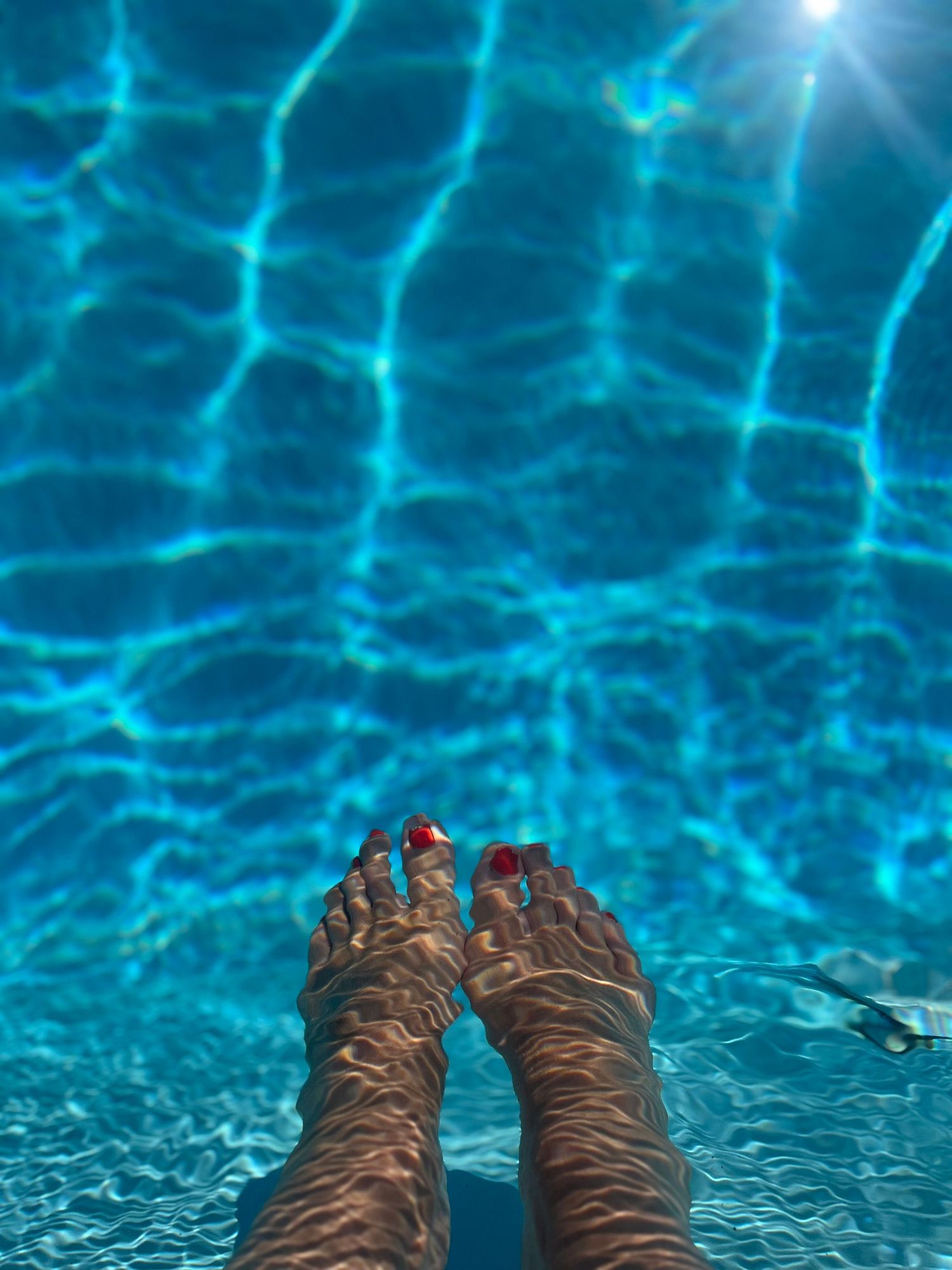 Feet with red toenail polish submerged in a bright blue swimming pool on a sunny day.
