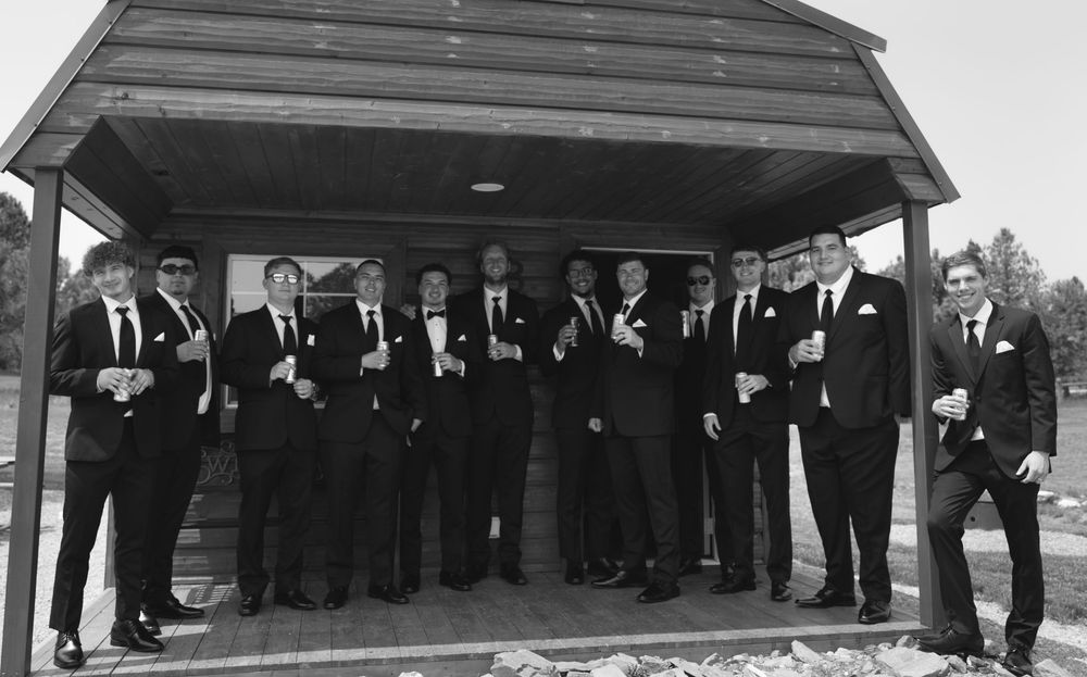 Group of groomsmen in black tuxedos posing together under a wooden gazebo in a black and white wedding photo.