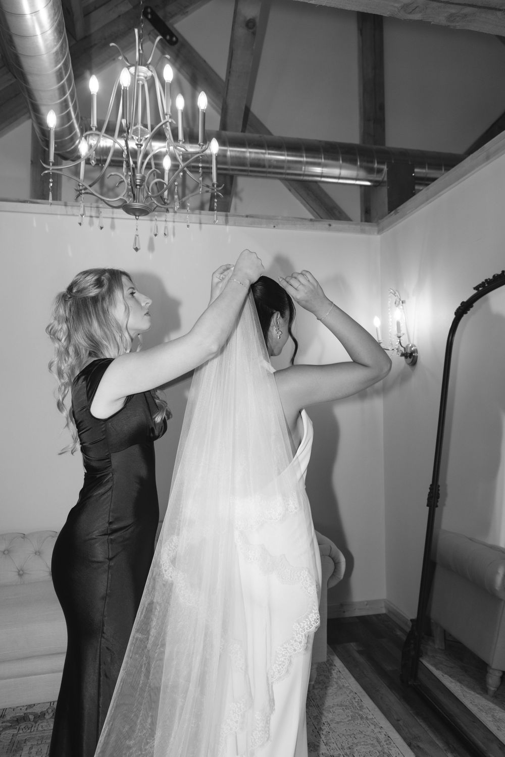 Bridesmaid in black dress helps bride adjust wedding veil in elegant room with chandelier, black and white photo.