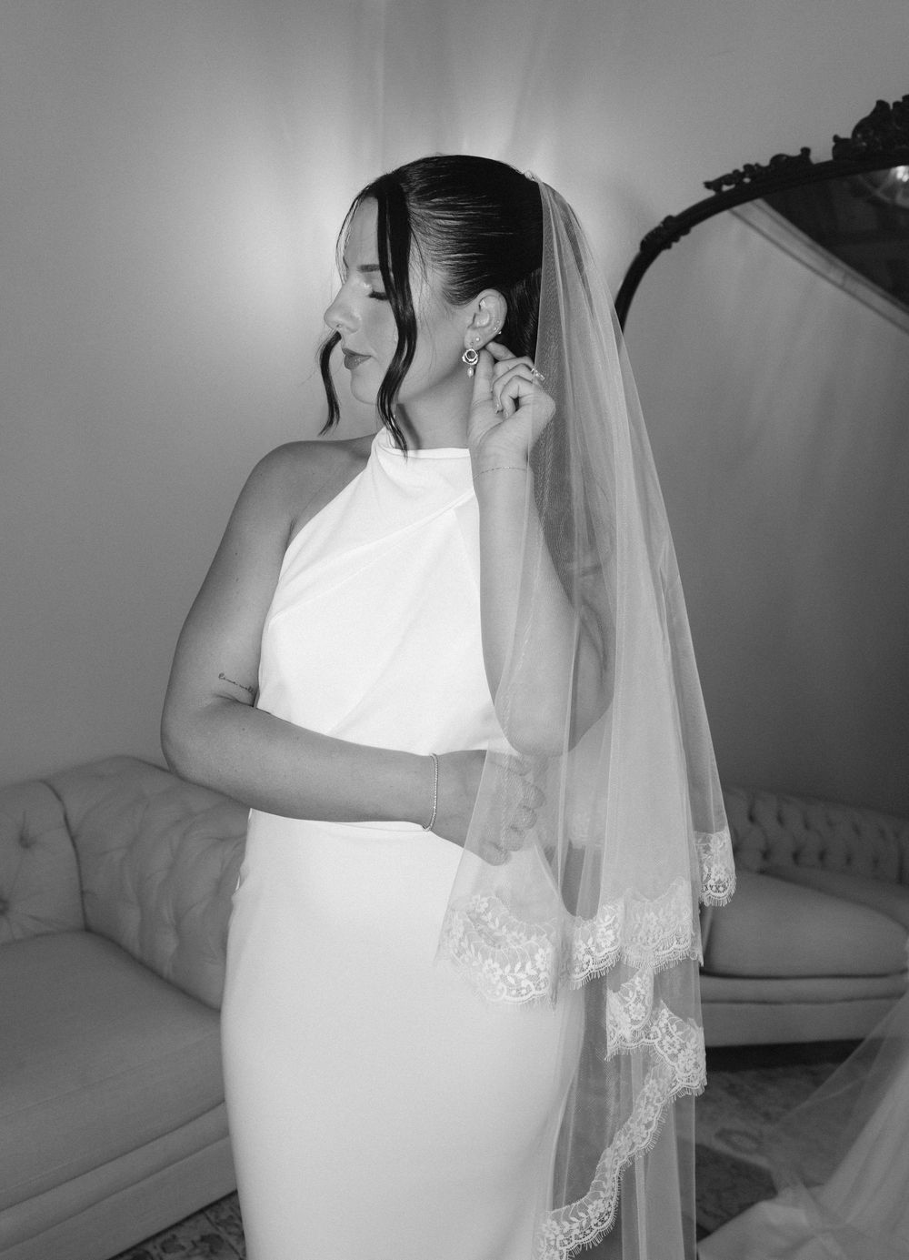 Black and white photo of a bride in a sleek halter gown and lace-trimmed veil adjusting her earring.