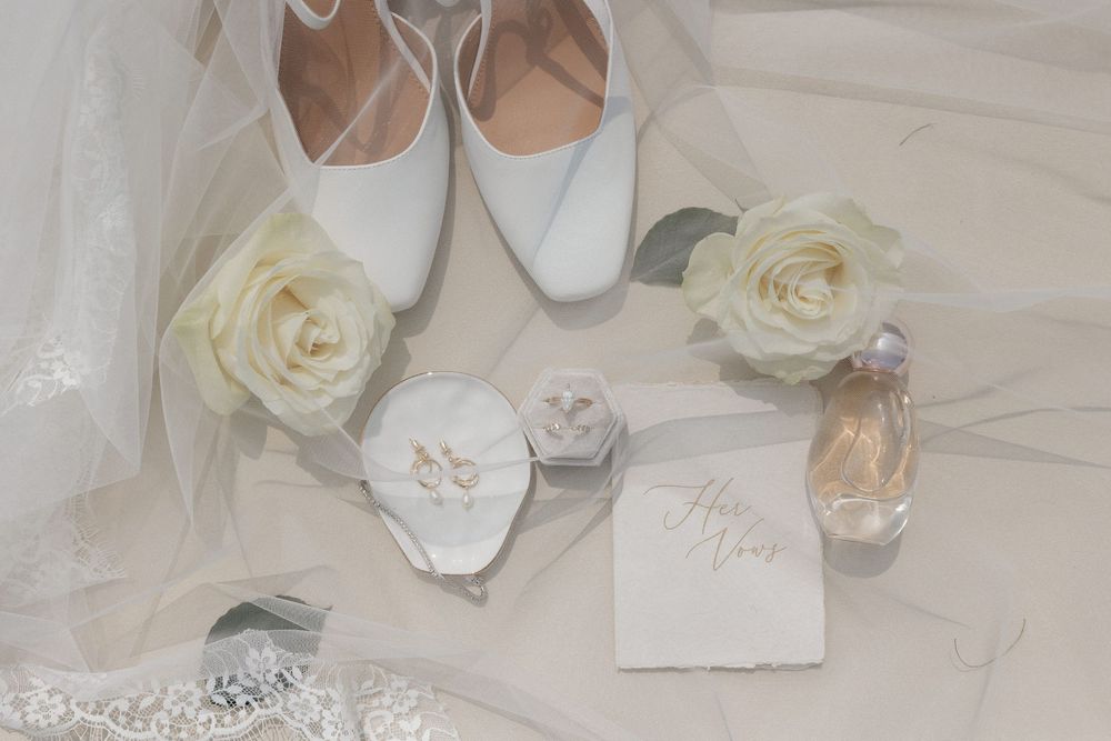 Elegant bridal flat lay with white heels, cream roses, wedding rings, veil, and perfume on a soft white background.