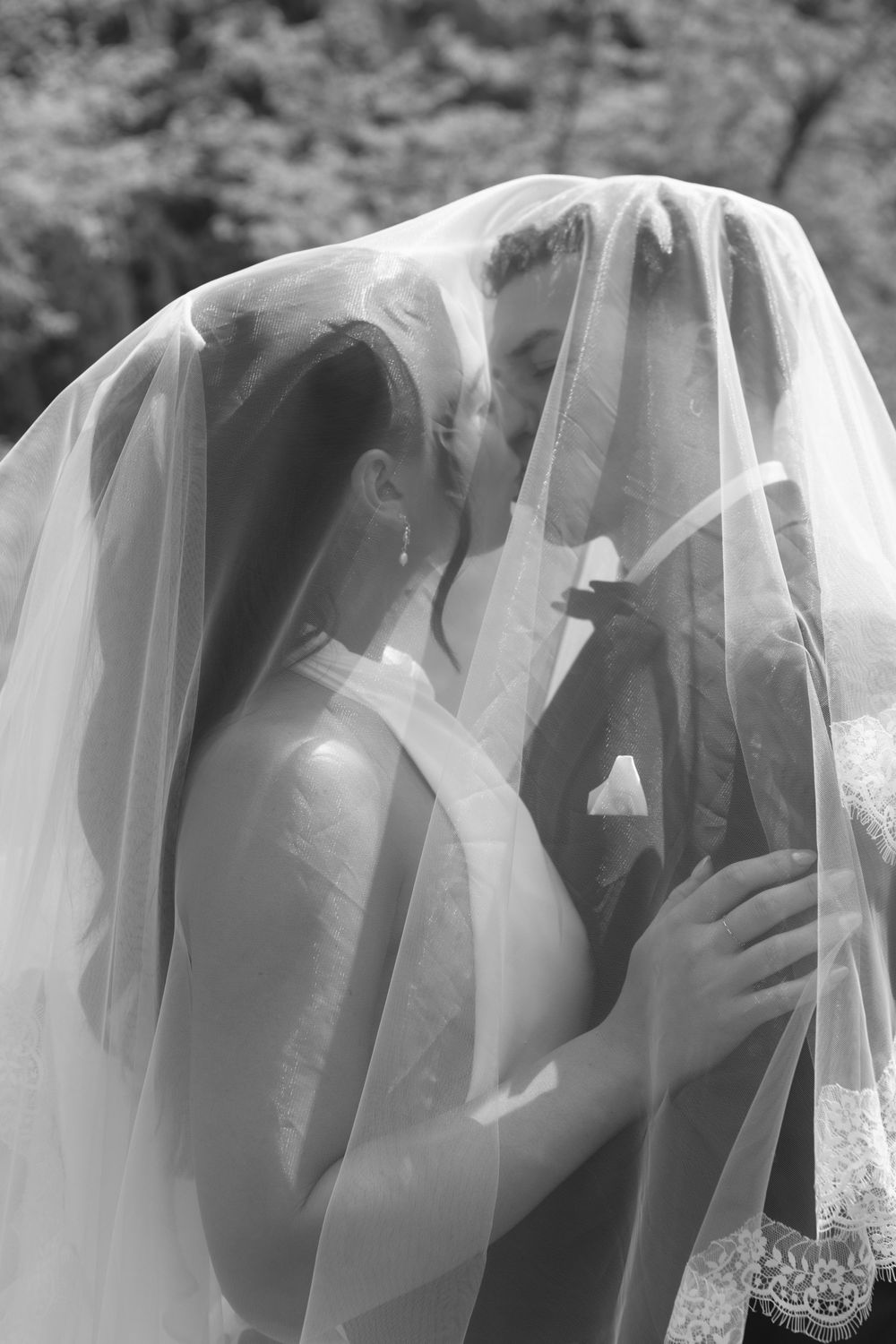 Black and white photo of bride and groom sharing a kiss under a flowing wedding veil outdoors.