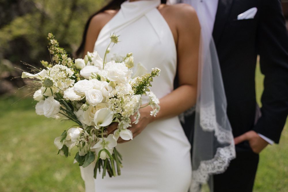 Bride holding a white floral bouquet with roses and greenery, standing beside groom in black suit outdoors.