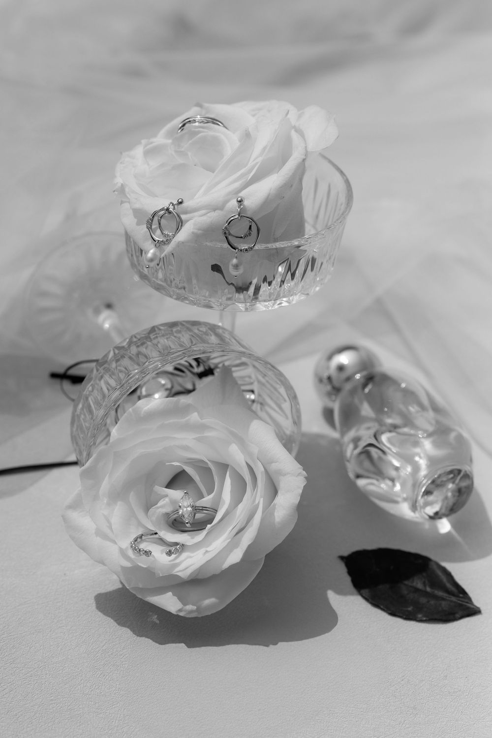 Black and white photo of white roses and crystal glasses with elegant silver earrings for a wedding flat lay.