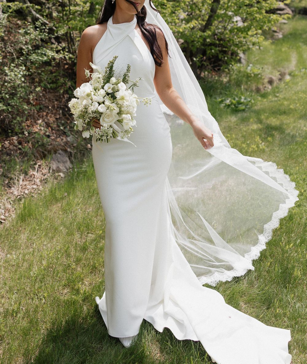 Bride in white halter mermaid wedding gown with flowing veil, holding white floral bouquet outdoors.
