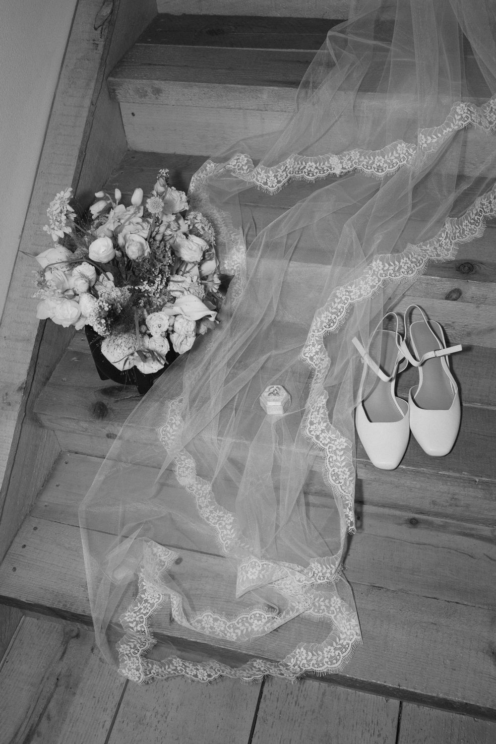 Black and white photo of bridal accessories on wooden stairs, featuring a lace veil, floral bouquet, heels, and ring.