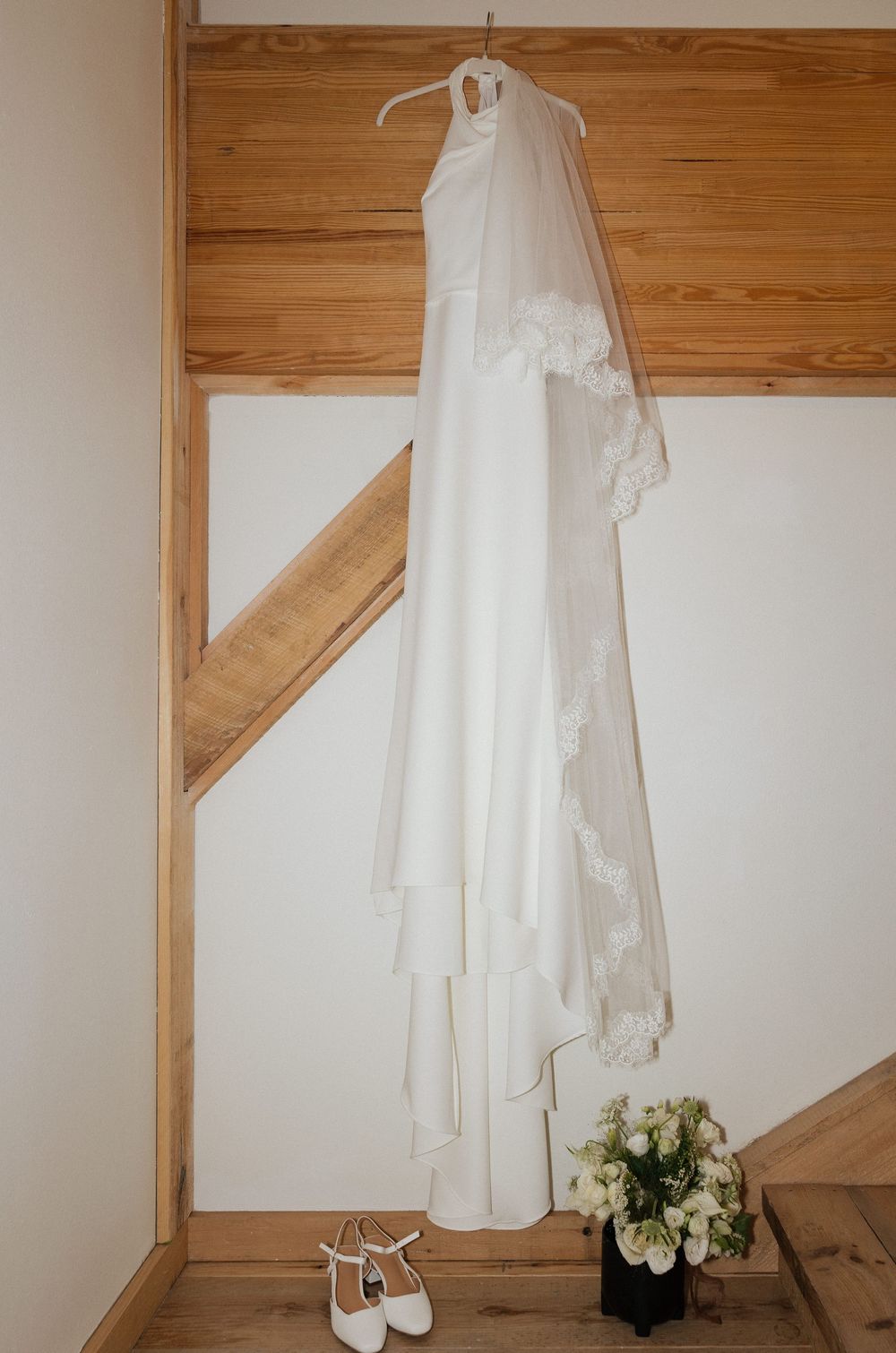 White halter wedding dress with lace-trimmed veil hanging in rustic barn, with bridal bouquet and white heels below.