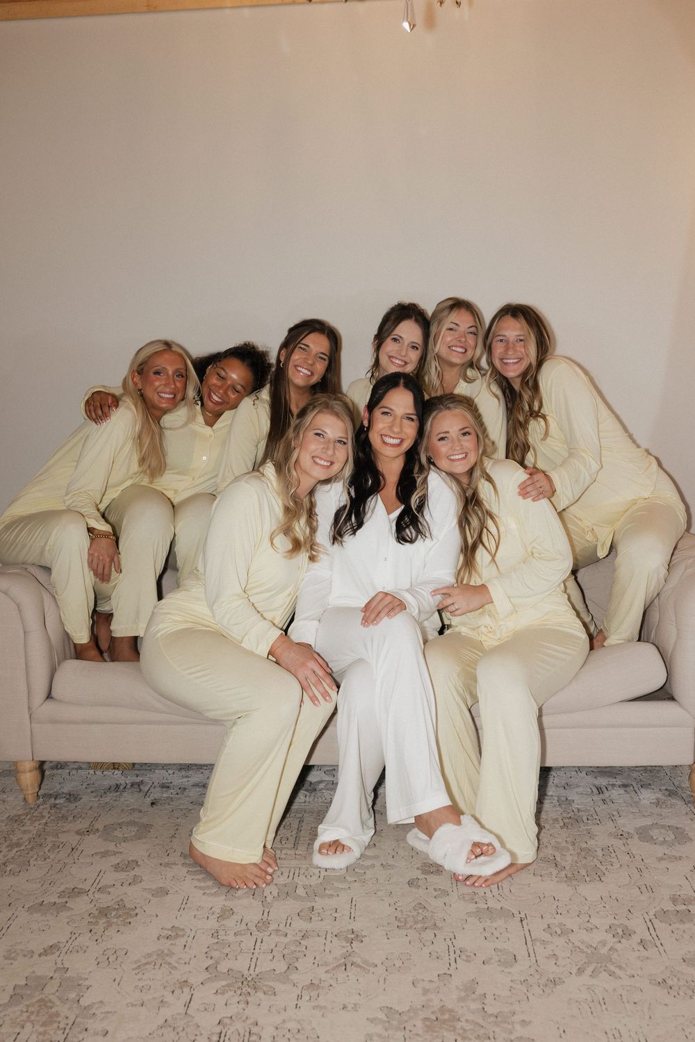 Bride in white robe poses with bridesmaids in matching cream pajamas on a couch during wedding morning prep.