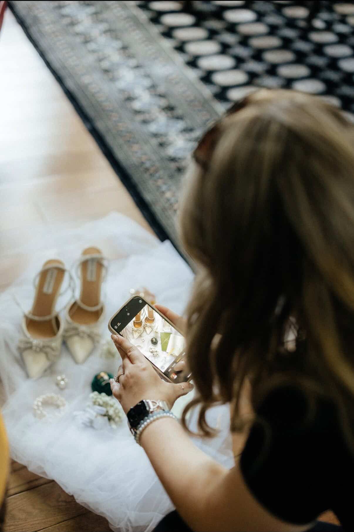 Woman photographing bridal accessories including heels and jewelry on white fabric with her smartphone.
