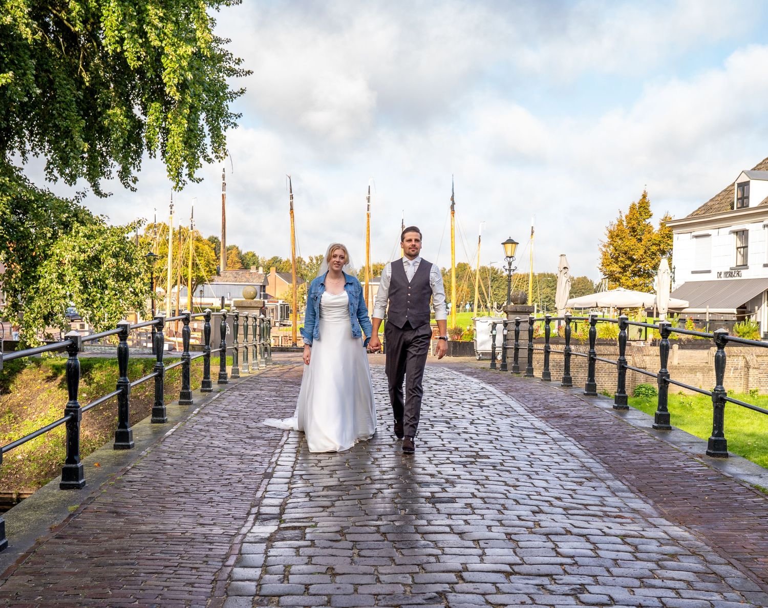 Bride and groom walk hand-in-hand on a cobblestone bridge near a harbor with sailboats on a cloudy day.