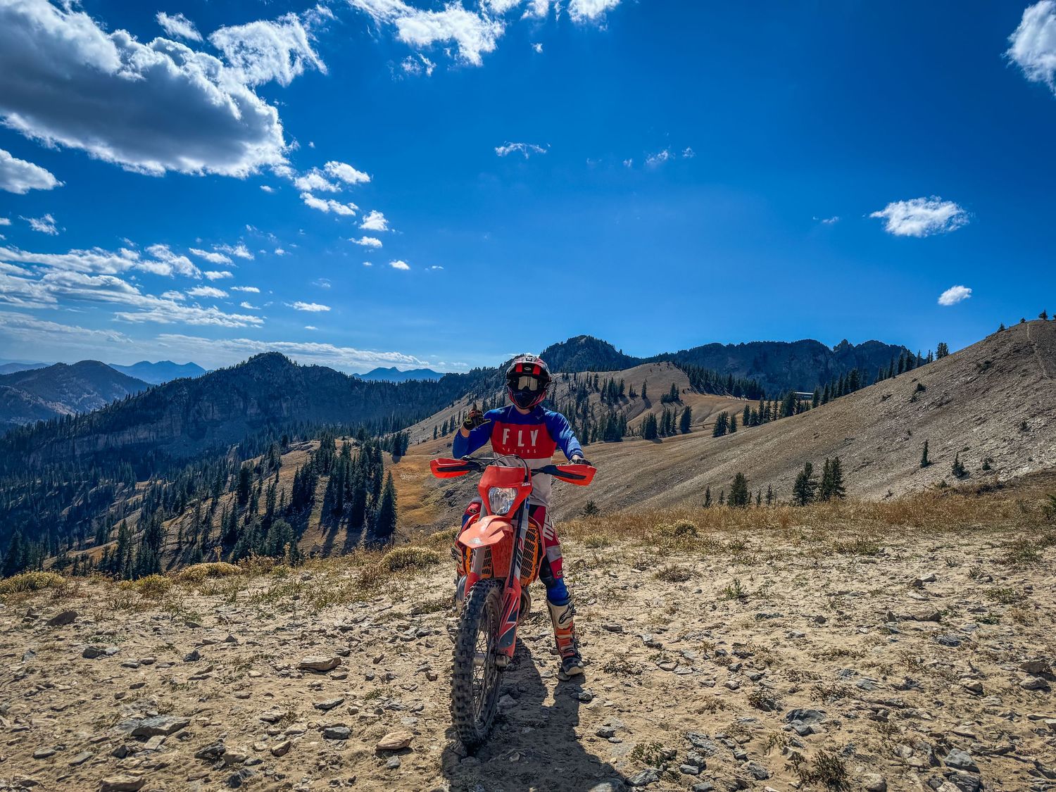 Motocross rider on a red dirt bike posed on a mountain ridge with scenic valley and blue sky backdrop. Captured by Slate Media (best in UT)