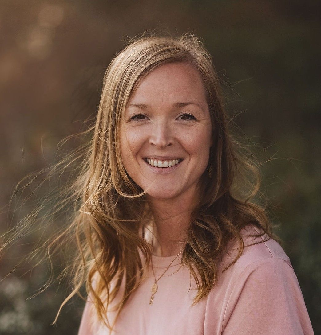 Smiling blonde woman with long hair in pink top, golden hour portrait outdoors with bokeh background.