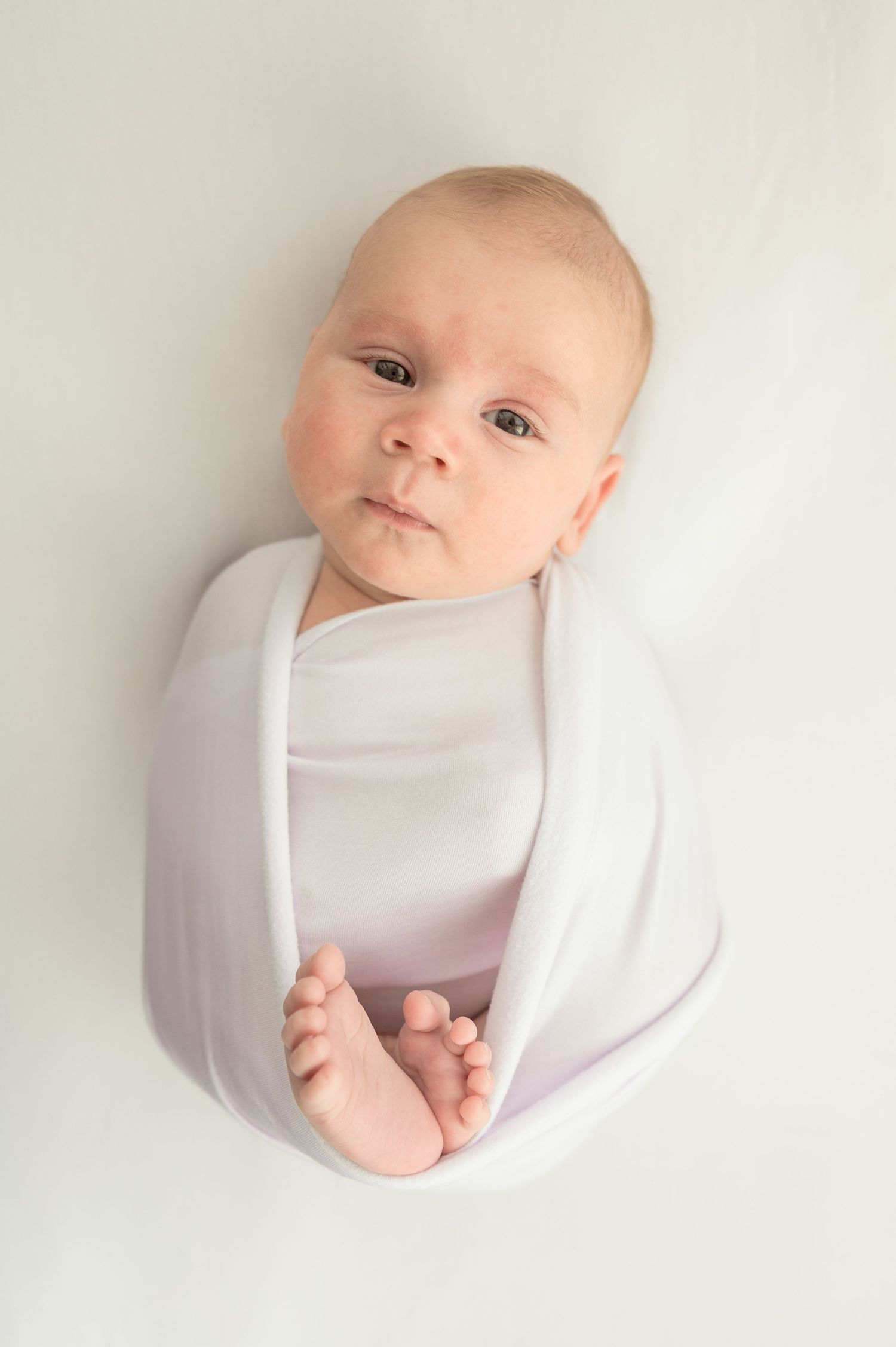 Newborn baby swaddled in a soft white wrap, lying on a white background with tiny feet peeking out.