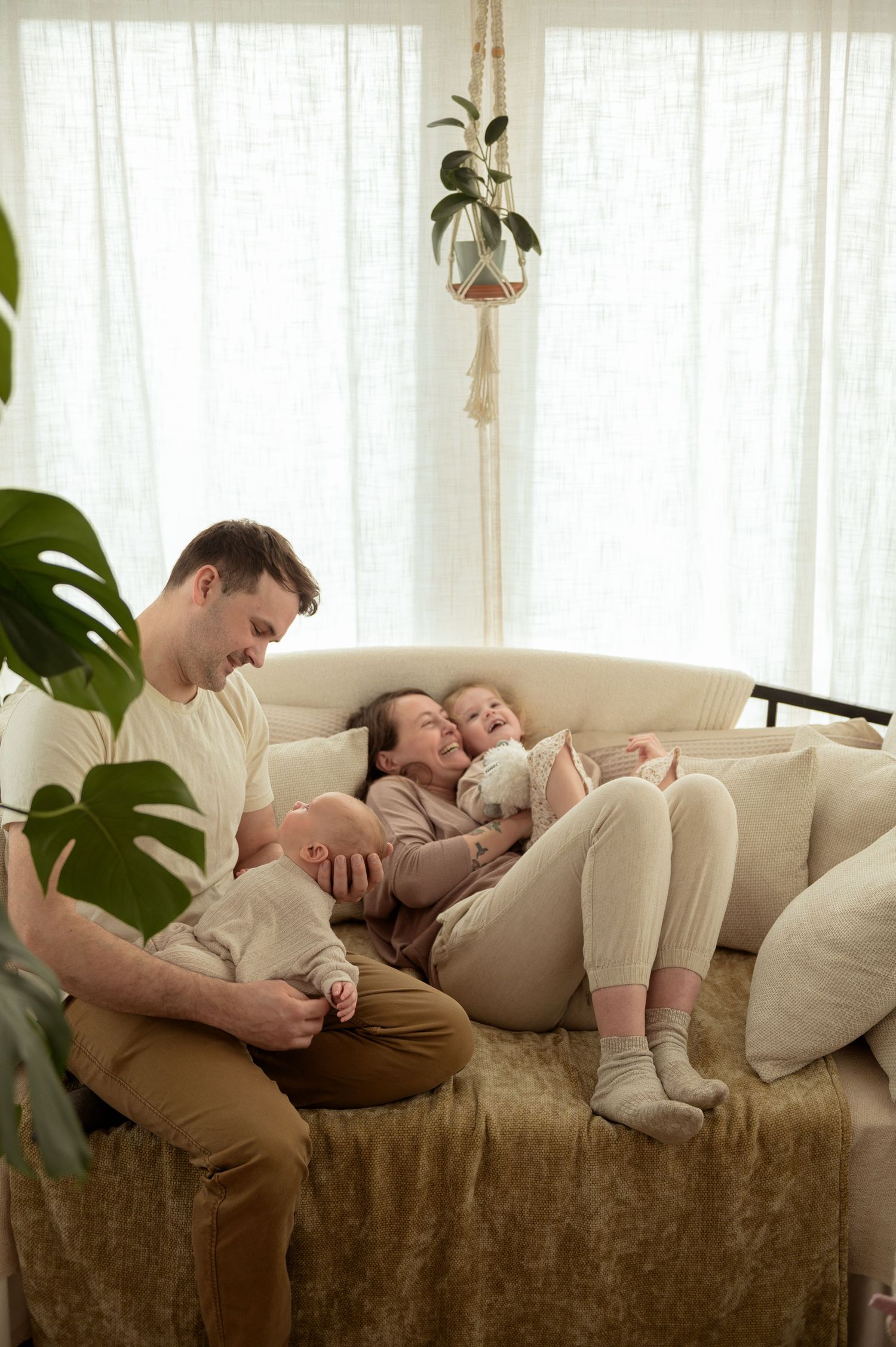 Happy family with two young children relaxing together on a cozy beige sofa in a bright, plant-filled living room.