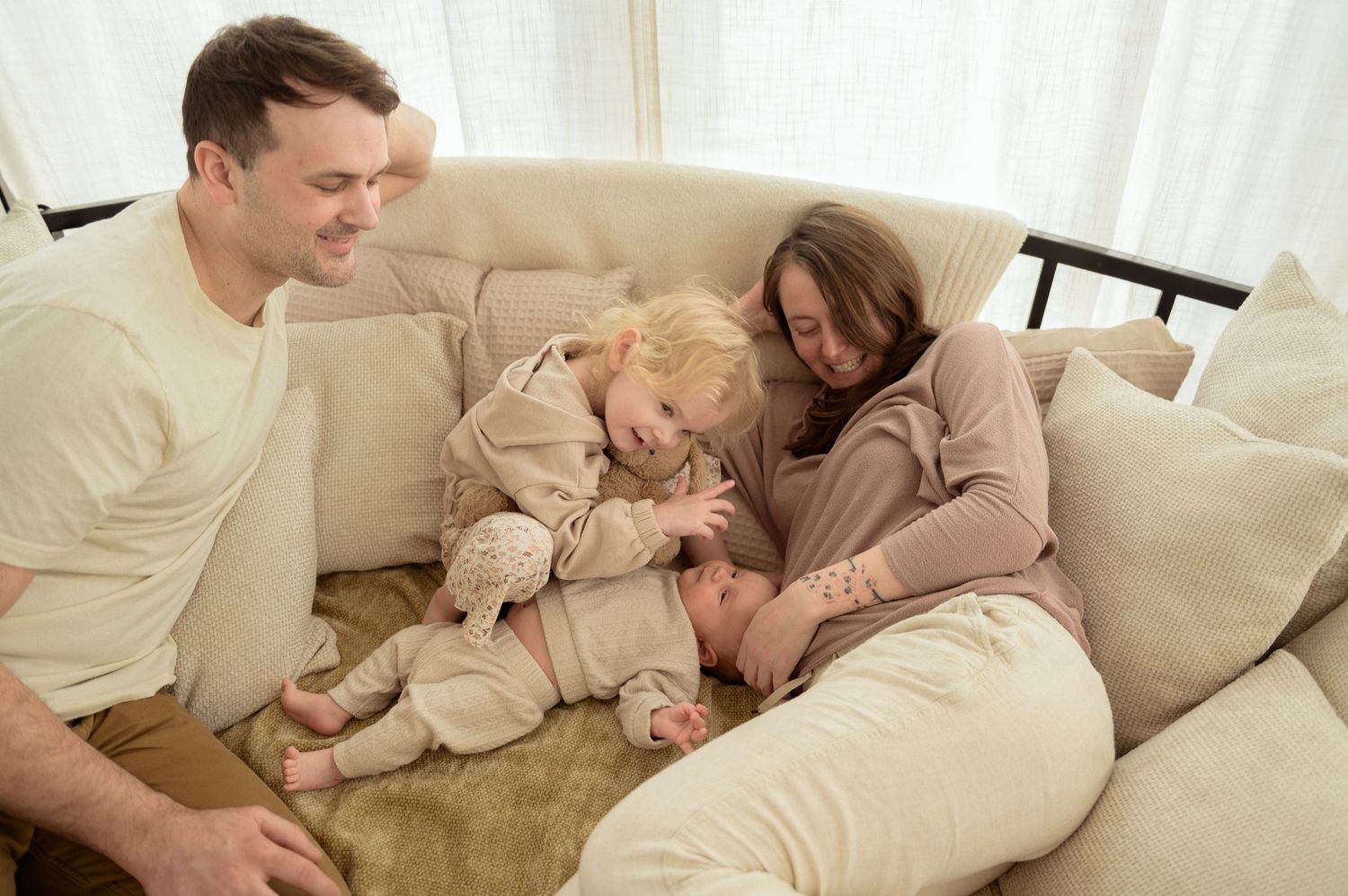 Happy family with toddler and newborn baby relaxing together on a cozy beige sofa at home.
