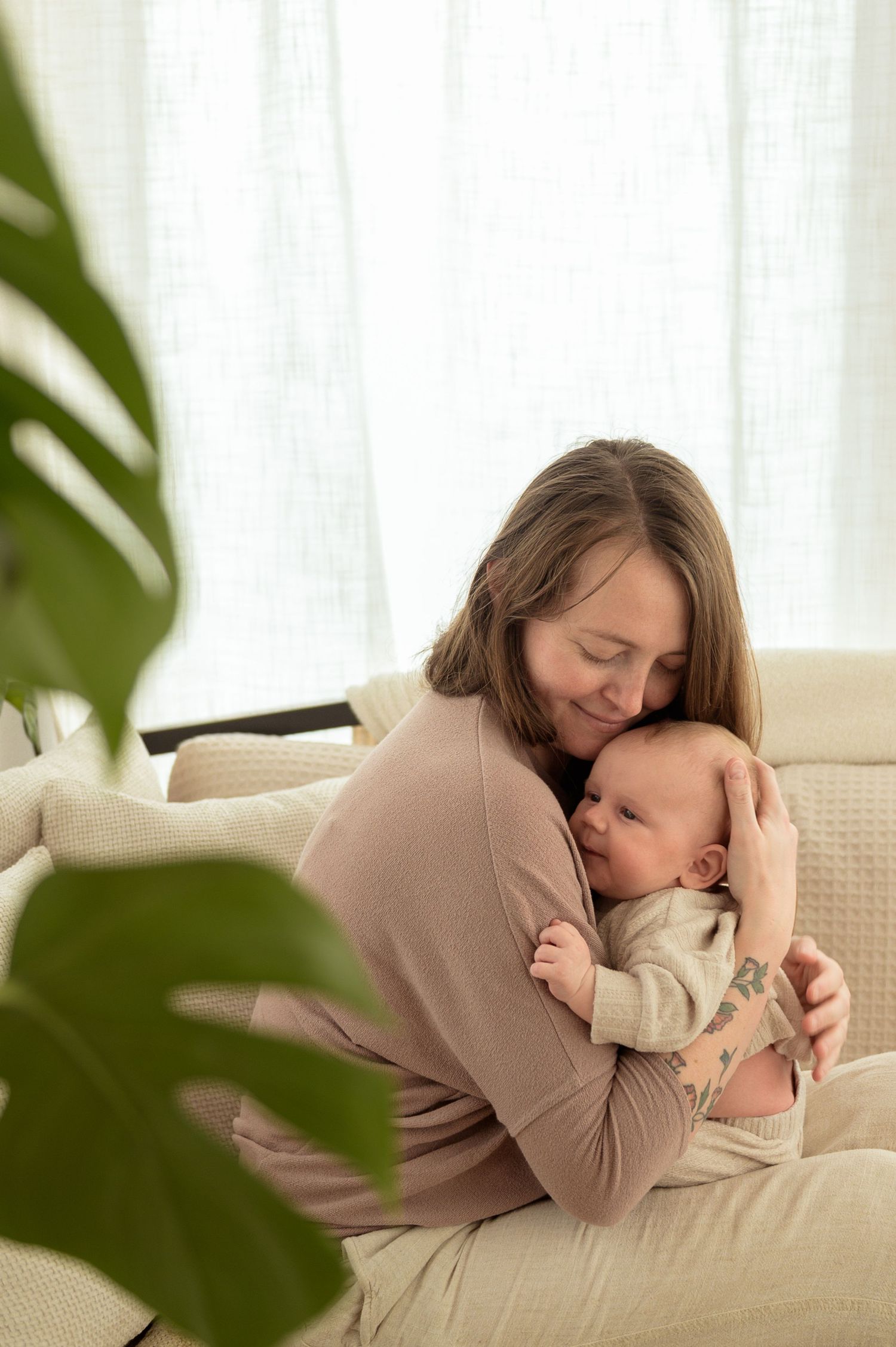 A mother tenderly embraces her newborn baby while sitting on a couch near a bright window.