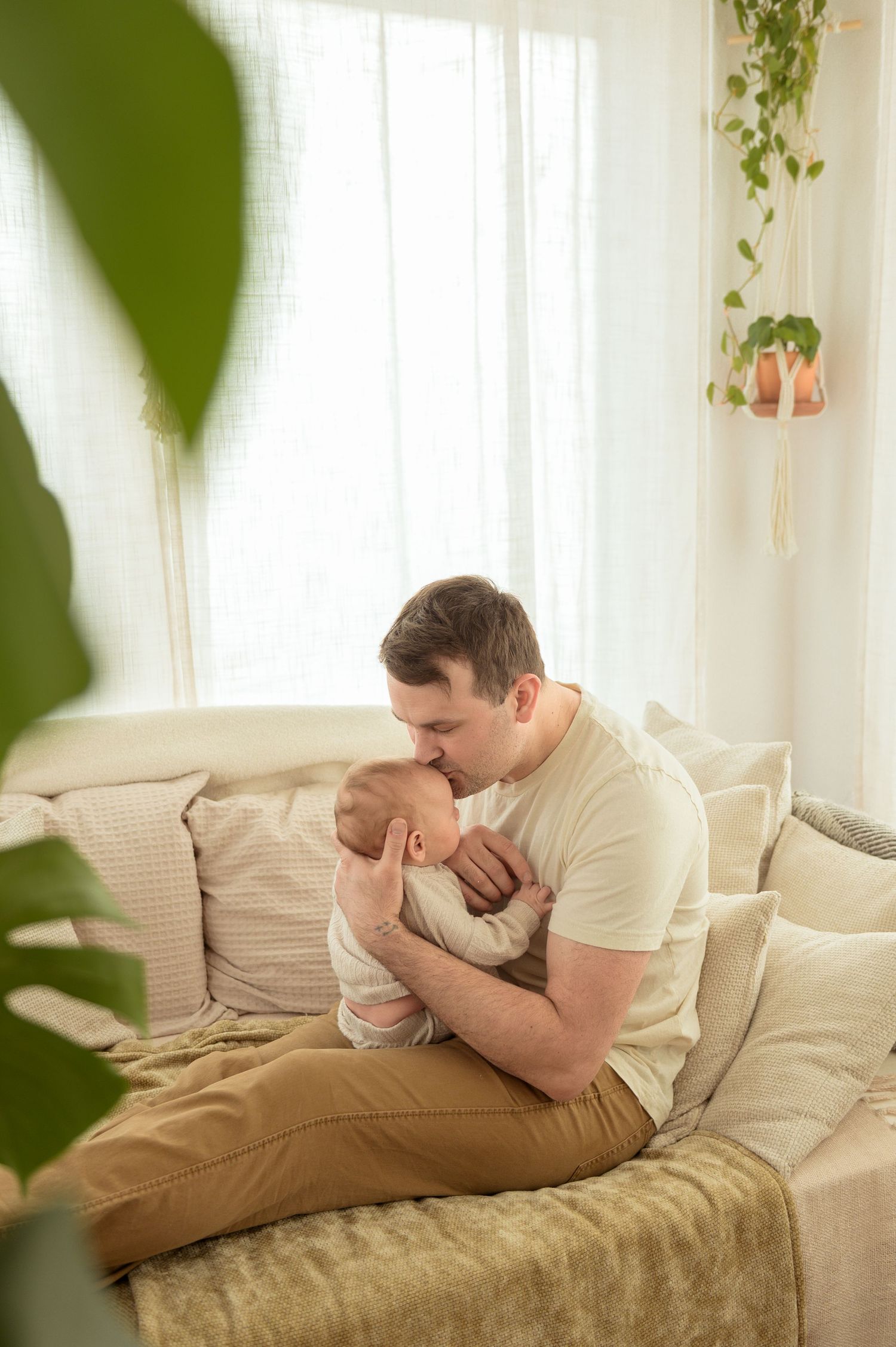 Father gently kissing his newborn baby while sitting on a cozy beige sofa in a bright, plant-filled living room.