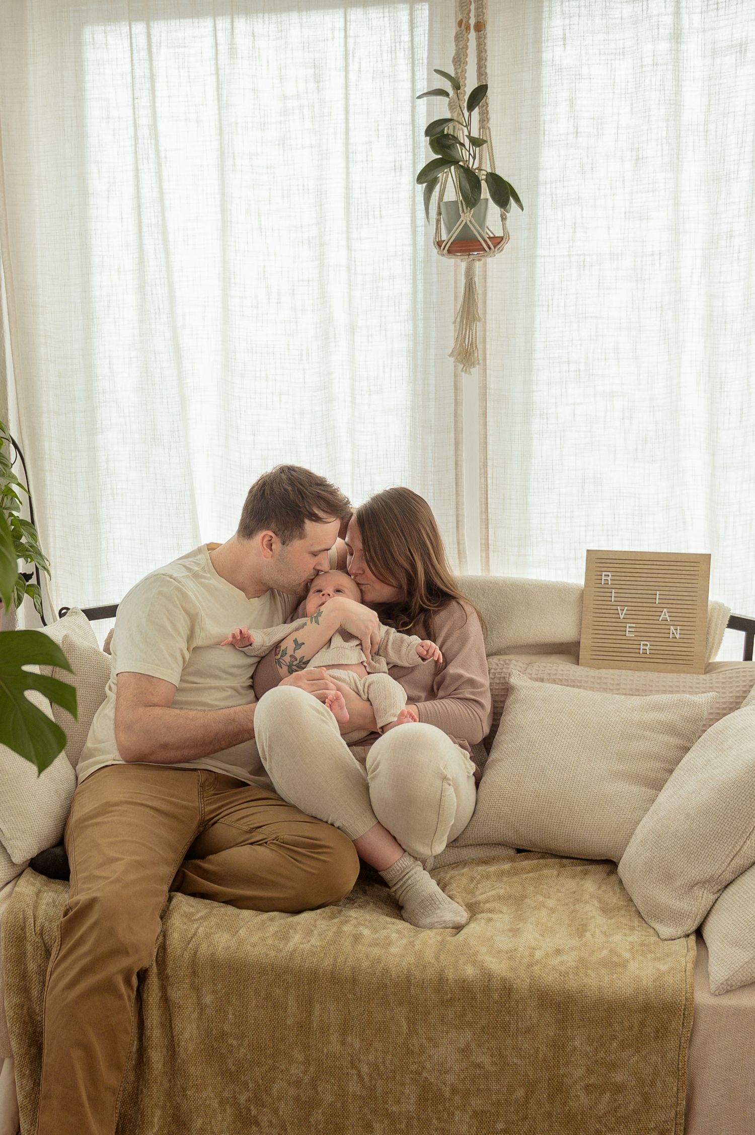 Happy parents sitting on a cozy sofa, lovingly gazing at their newborn baby in a bright, plant-decorated living room.