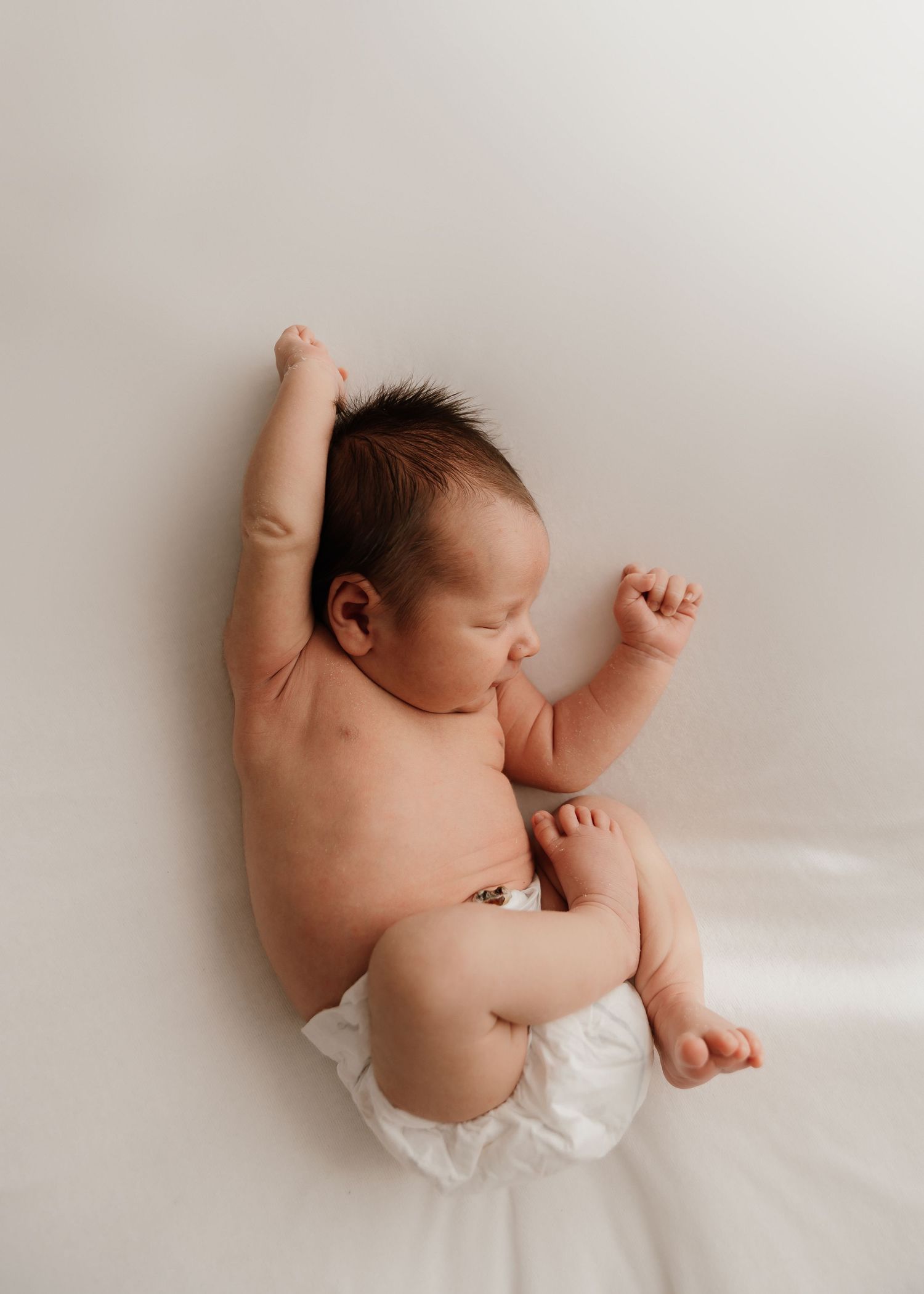 A sleeping infant dressed in white lies on a soft white surface in a series of gentle stretching poses.