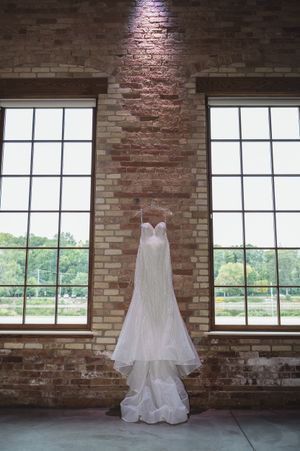 A white wedding dress hangs elegantly in front of large windows in a rustic brick loft space.