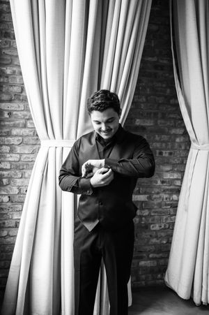 Black and white photo of a person in formal attire checking their phone against a brick wall with white curtains.