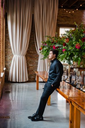 Bartender in dark uniform demonstrates mixing drinks at a rustic venue with exposed brick walls and white curtains.