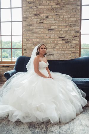 A bride in a voluminous white wedding gown lounges gracefully on a navy blue couch against an exposed brick wall.