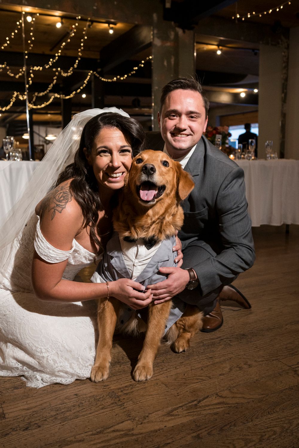 A happy golden retriever poses with a couple during their wedding celebration on a wooden floor under string lights.