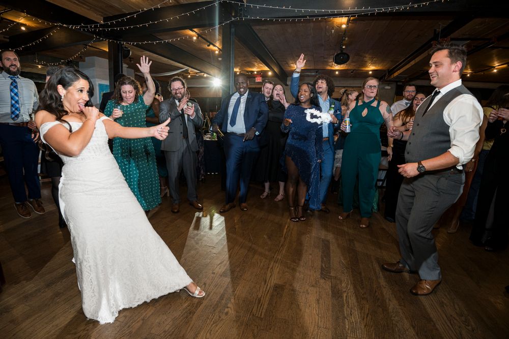 Wedding reception guests dance and celebrate on a rustic wooden dance floor under warm lighting and string lights.