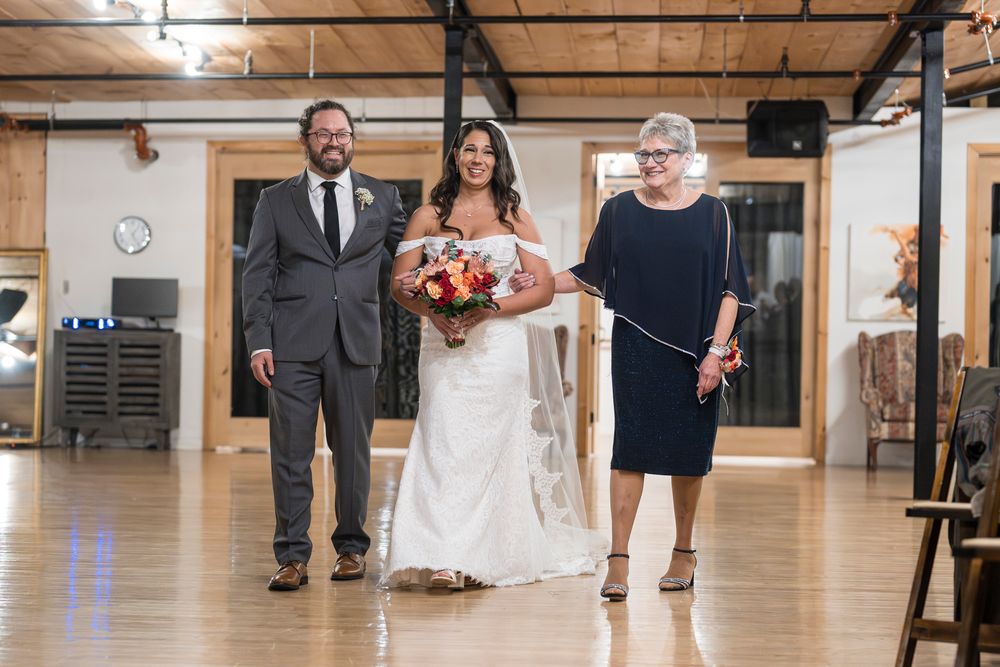 A bride in a white lace gown holds a coral bouquet while walking down a wooden dance floor with two companions.