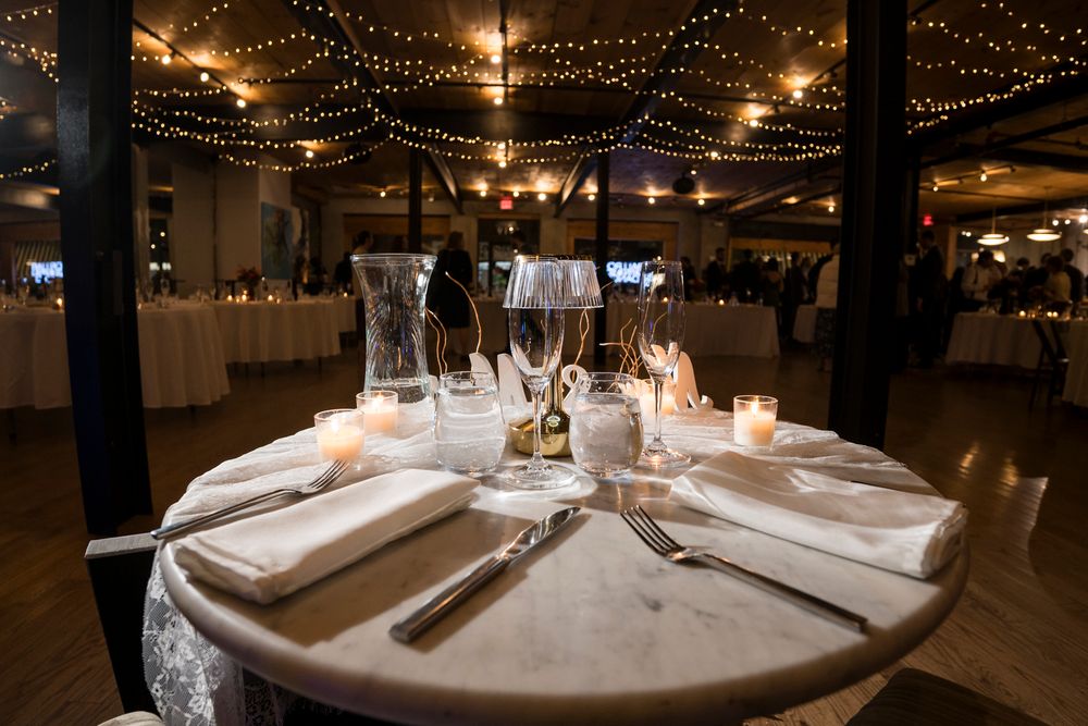 Wedding reception tables elegantly set with white linens and glowing candles under romantic string light decorations.