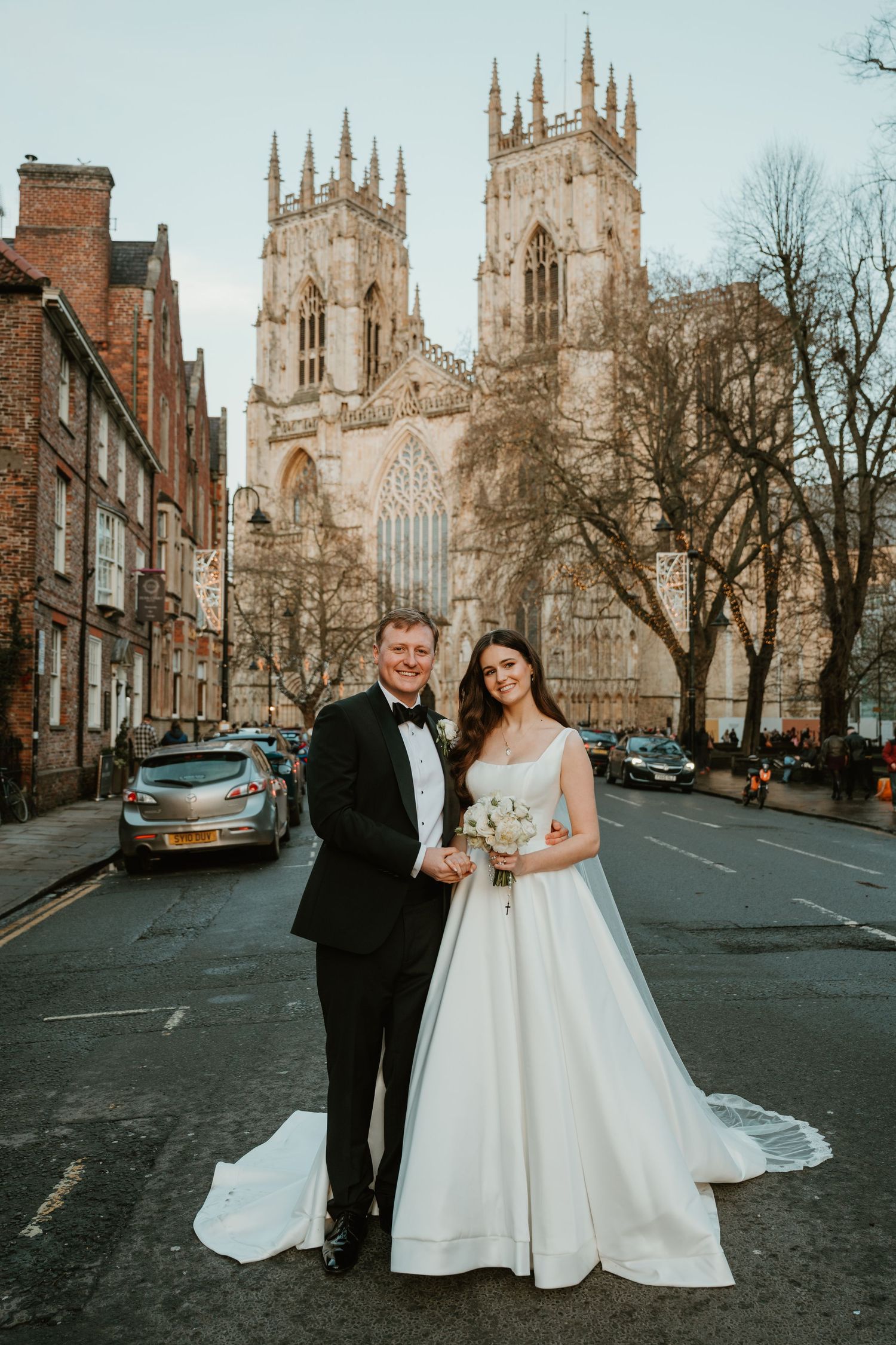 A couple poses for wedding photos outside York Minster cathedral on a winter afternoon with bare trees in the background.