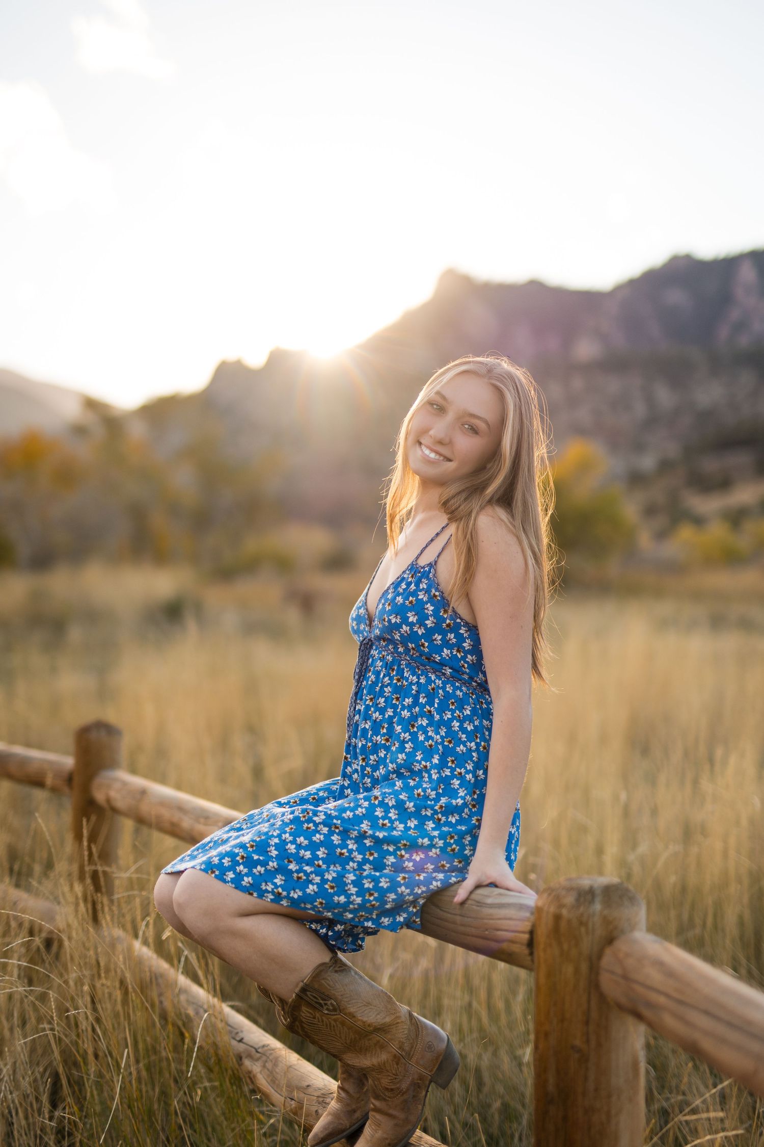 A person in a blue floral dress sits on a rustic wooden fence at sunset in a golden meadow.