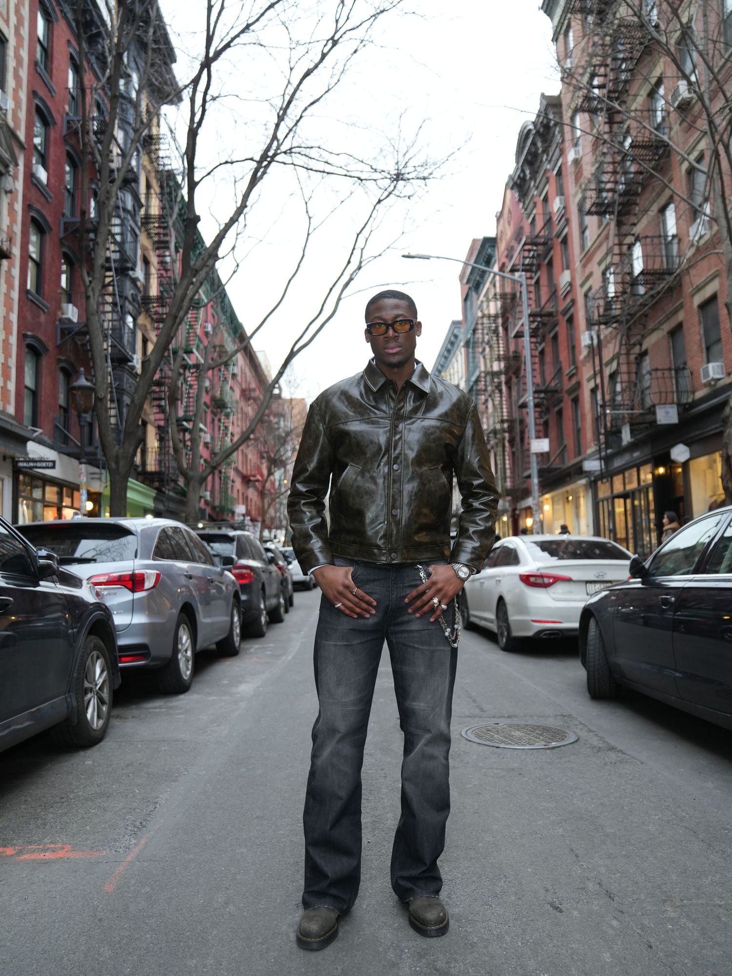 Tall man in dark leather jacket standing confidently on a New York City street lined with parked cars.