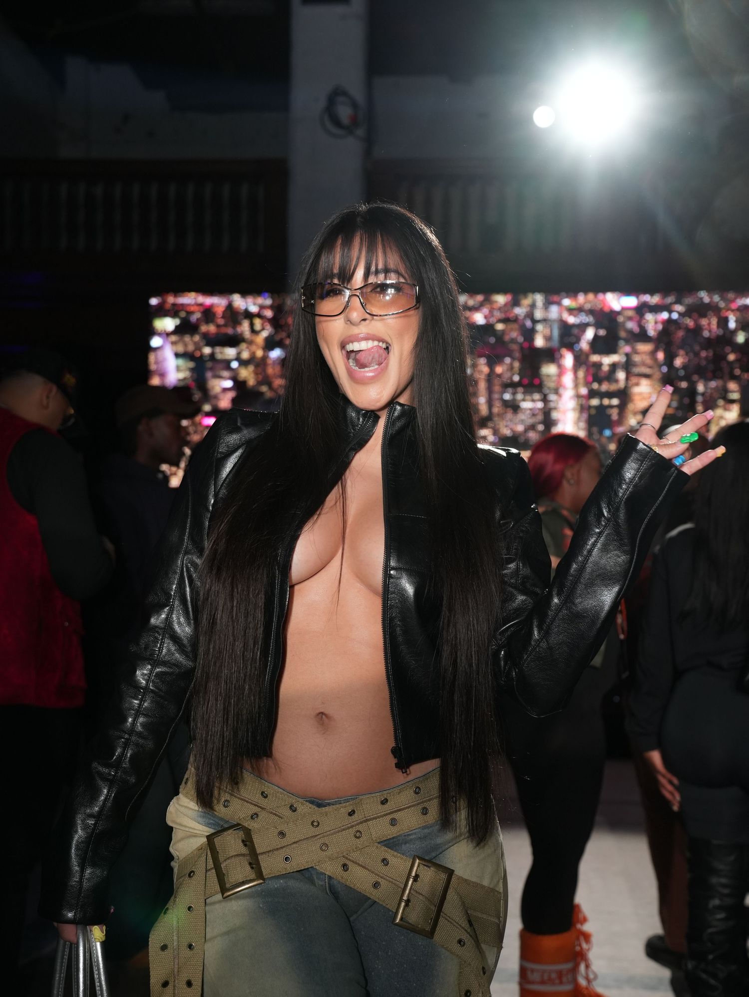 Woman in black leather jacket with open front posing energetically at a nightclub event with city backdrop.