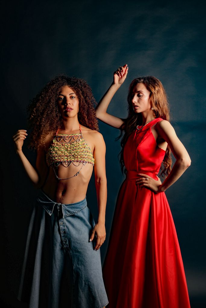 Two women posing in studio, one in gold chain crop top with denim skirt, other in red gown.