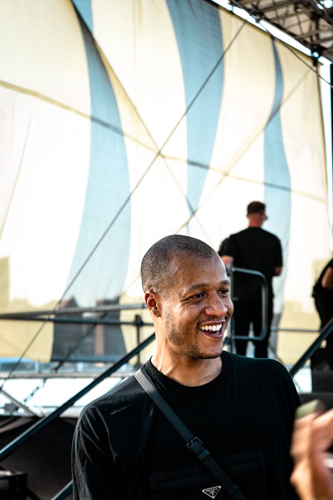 Smiling young Black man in black Prada shirt laughing outdoors at a New York fashion event.