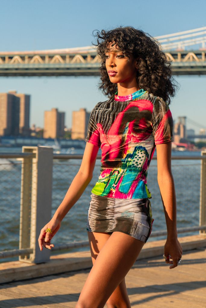 Fashion model in colorful graphic mini dress posing on waterfront with Manhattan Bridge backdrop.