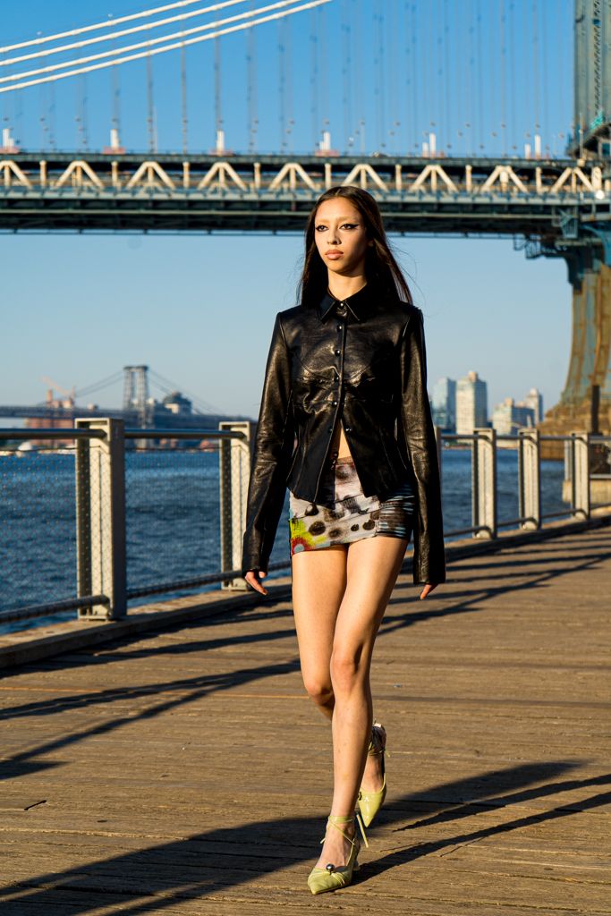 Model in black leather jacket and mini skirt walking on Brooklyn waterfront boardwalk near bridge.