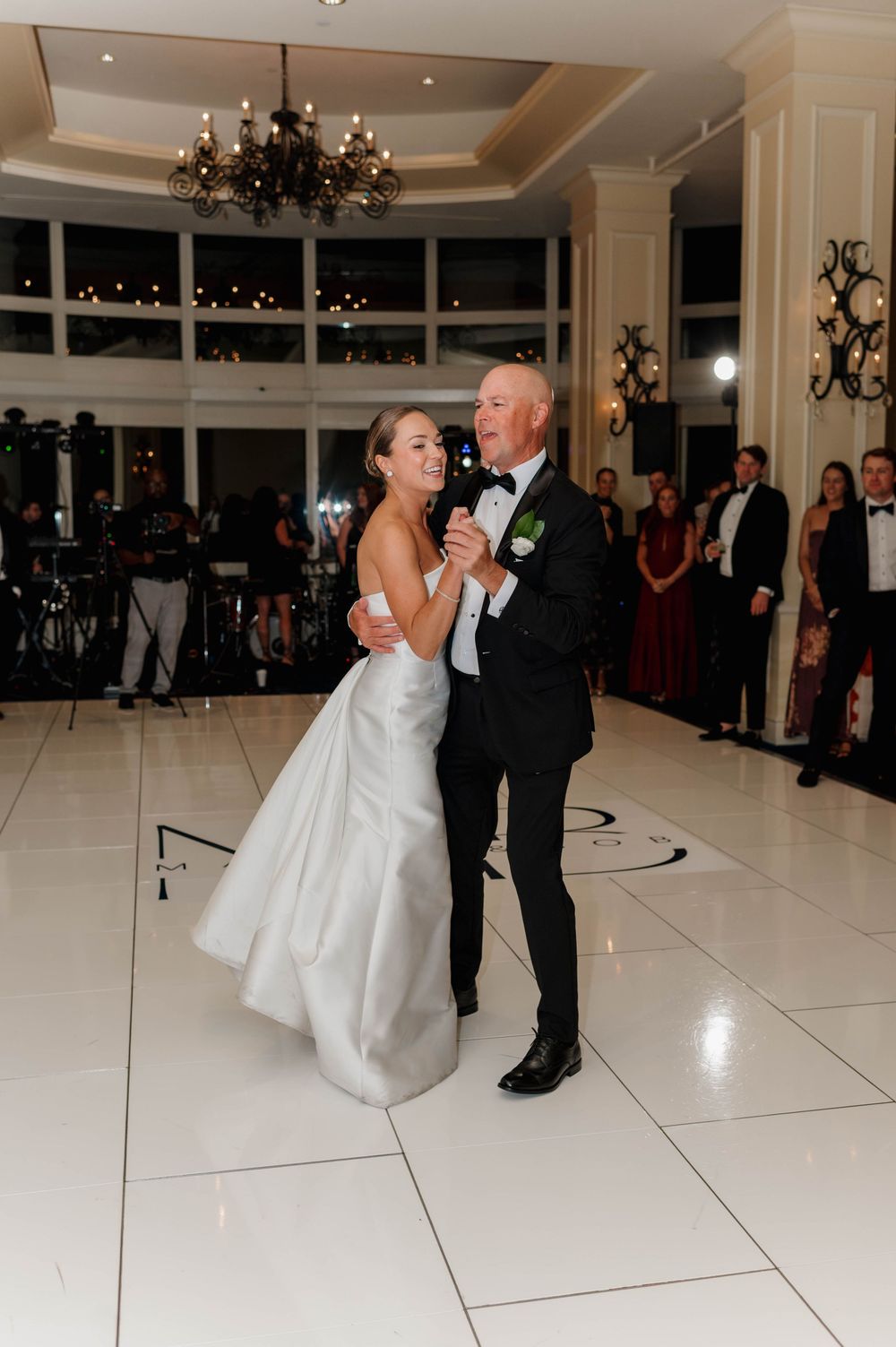 A couple shares their first dance in an elegant white ballroom with chandeliers and large windows.