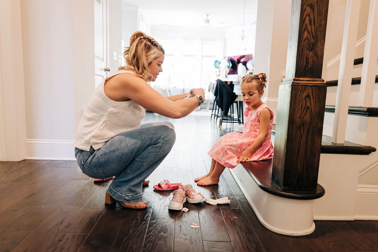 A little girl sits on a staircase with her mom helping her put her shoes on.