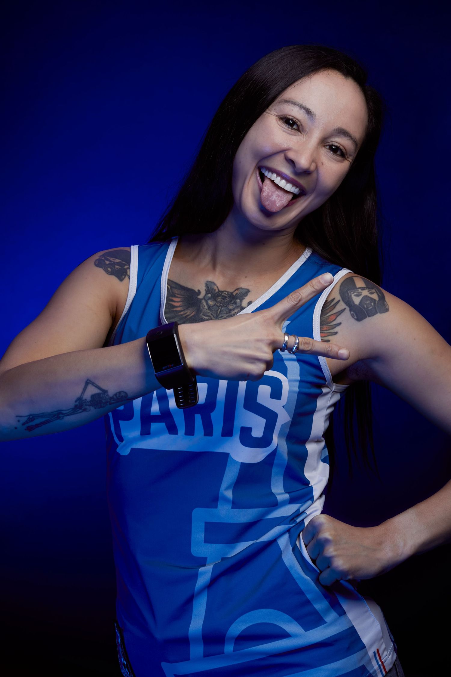 Woman in blue Paris jersey sticking tongue out, showing tattoos and smartwatch, posing playfully on blue background.