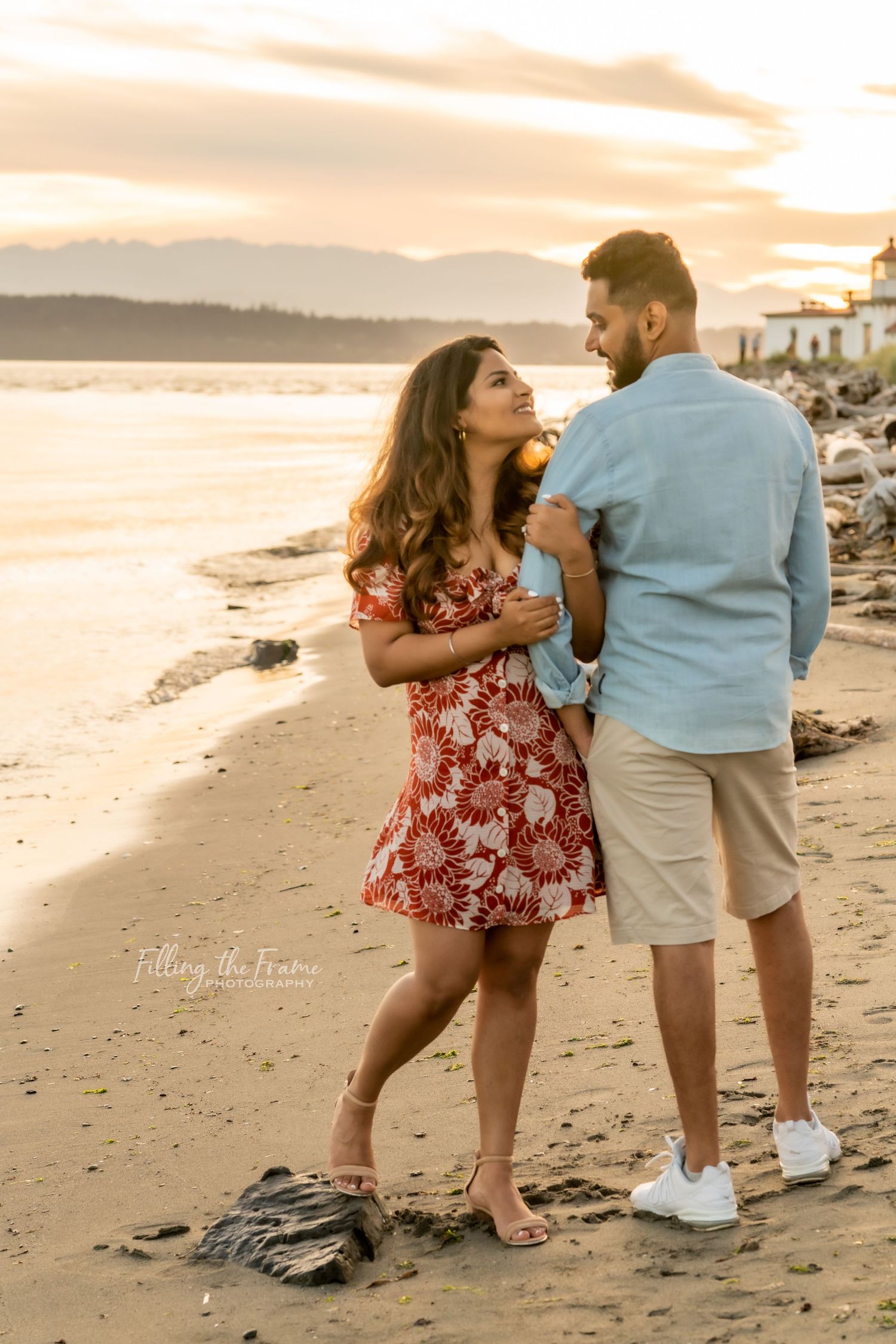 Couple embracing on a sandy beach at golden sunset, woman in red floral dress, man in light blue shirt and khaki shorts.