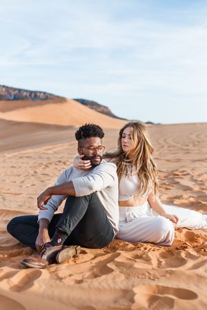 A couple sits together on golden sand dunes in a desert landscape during sunset.