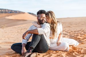 A romantic couple shares intimate moments while sitting on red desert sand with mountains in the distance.