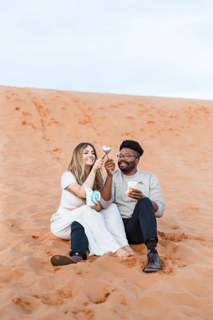 A couple laughs together while sitting on orange desert sand during a photo session.